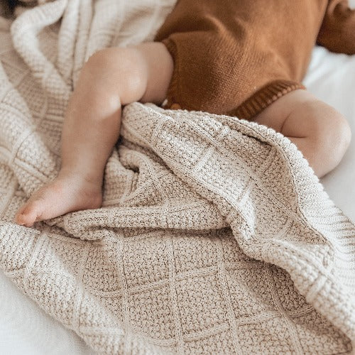 Baby laying on a bed with a brown onsie on and covered in a Snuggly Jacks Taupe Knitted Blanket